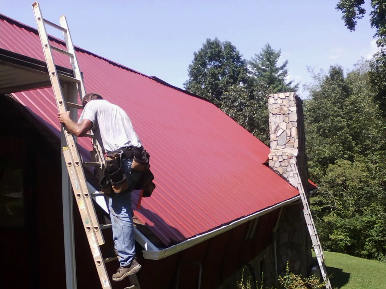 Roofer on a ladder beside a red metal roof and stone chimney — Soffit & Fascia Repair by Roofing Brothers in Millburn